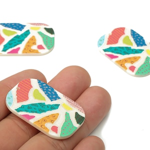 May include: Rectangular, terrazzo-style resin cabochons in a variety of colors. The cabochons feature a white base with colorful abstract shapes in pink, orange, blue, green, and yellow. One cabochon is held in a hand, with two others nearby.
