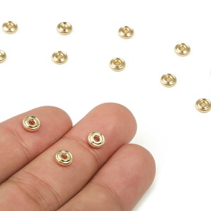 May include: A collection of small, gold-colored metal beads with a circular design and a central hole. Several beads are scattered on a white surface, with three beads resting on a person's fingers. These beads are likely used for jewelry making.