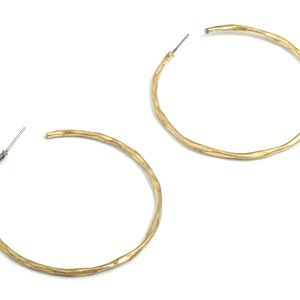 May include: Two gold-colored hoop earrings with a textured, hammered appearance. The earrings are circular, with a simple post closure. The metal has a slightly irregular surface, giving them a handcrafted look. The earrings are displayed against a white background.