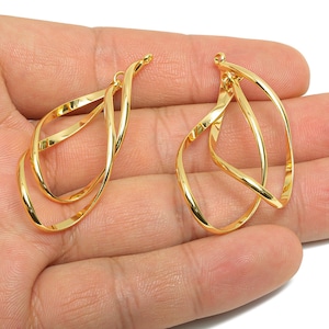 May include: Two gold-colored, abstract-shaped earrings. Each earring features a looping, intertwined design. The earrings are displayed against a neutral background, highlighting their polished, reflective surface. The jewelry is designed for pierced ears.