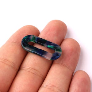 Peut inclure: Un maillon ovale translucide dans les tons de bleu, vert et blanc. Le maillon est tenu dans une main sur fond blanc. Ce maillon décoratif pourrait être utilisé pour la fabrication de bijoux ou d'autres projets d'artisanat.