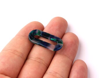 May include: An oval-shaped, translucent link in shades of blue, green, and white. The link is held in a hand against a white background. This decorative link could be used for jewelry making or other craft projects.