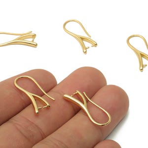 May include: Gold-colored earring findings with a hook design. The findings have a V-shaped base with a small setting, and a curved hook for attaching to the ear. Several pieces are scattered on a white background.