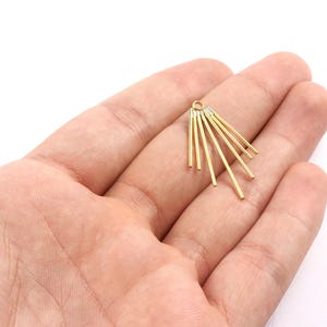 Könnte beinhalten: Ein goldfarbener Metallanhänger mit einem fächerartigen Design. Der Anhänger hat mehrere dünne, gerade Stäbe, die von einem zentralen Punkt nach außen strahlen. Der Anhänger wird in der Hand einer Person vor weißem Hintergrund gehalten.