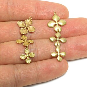 Può includere: Due serie di ciondoli floreali color oro. Ogni ciondolo presenta tre o quattro pendenti a forma di fiore collegati da piccoli anelli. I ciondoli sono tenuti in mano su sfondo bianco. Questi ciondoli sono ideali per la creazione di gioielli.