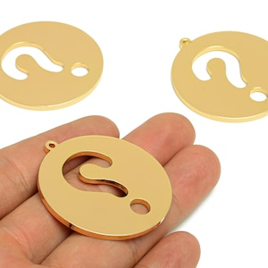 May include: Gold-colored metal pendants in a circular shape, each featuring a cutout question mark design. The pendants have a small hole at the top for stringing. One pendant is held in a hand, with two others visible in the background.