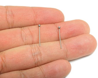 May include: Two silver-colored nose studs, each with a small ball at the top, are displayed. The studs are straight and thin, designed for piercing jewelry. The background is a light skin tone.