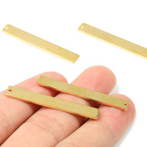 May include: Close-up of several gold-colored rectangular metal bar charms. Each charm has a small hole at one end. The charms are displayed against a white background.