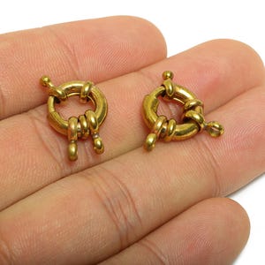 Puede incluir: Dos cierres de joyería redondos de color dorado. Cada cierre tiene un cuerpo circular con una abertura con bisagras y un pequeño botón decorativo. Los cierres se sostienen en una mano y están listos para la fabricación de joyas.