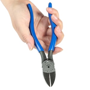 May include: Blue-handled pliers with black cutting edges. The pliers are held in a hand against a white background. The tool has the text "KEIBA JAPAN PL-717" printed on the metal.