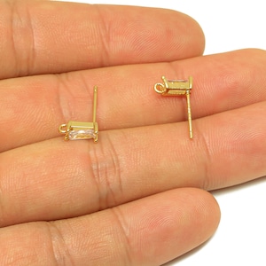 May include: Two gold-colored stud earrings with rectangular, clear gemstones. The earrings are held in a hand against a white background. The earrings have a post and a small loop for attaching a backing.