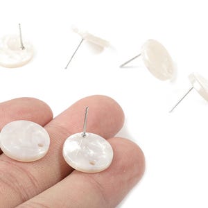 May include: White, round stud earrings with a pearlescent finish. The earrings have a post for pierced ears. Several earrings are scattered on a white background.