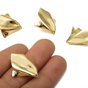 May include: Gold-colored, abstract stud earrings. The earrings have a unique, folded design with a shiny, reflective surface. The image shows the earrings on a white background, with one held in a hand.