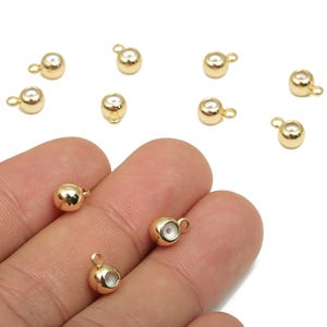 Könnte beinhalten: Goldfarbene Schmuckzubehörteile, darunter mehrere kleine, runde Perlen mit einer Schlaufe oben. Einige Perlen werden in einer Hand gehalten, um ihre Größe und ihr Design zu zeigen. Die Perlen haben einen kleinen, weißen Einsatz.