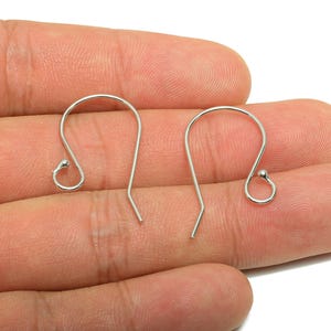 May include: Two silver earring hooks are shown. Each hook has a curved top with a small ball and a straight, angled bottom. The hooks are designed for jewelry making. The background is light.