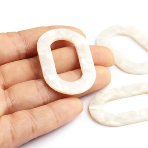 Peut inclure: Gros plan sur trois maillons ovales en résine blanc cassé. L'un est tenu dans une main, deux autres sont à proximité. Les maillons ont un aspect nacré et marbré, sur fond blanc. Idéal pour la création de bijoux.