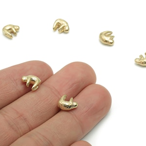 May include: Small, gold-coloured sloth charms. The charms are in various poses, with a textured surface. Several charms are scattered on a white background, with a hand holding two of the charms.