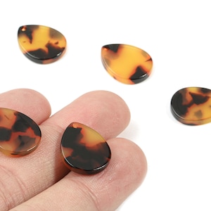 Può includere: Cinque perline a forma di goccia con un motivo tartarugato marrone e giallo. Due perline sono tenute in mano, mentre le altre sono sparse su una superficie bianca. Ogni perlina ha un piccolo foro.