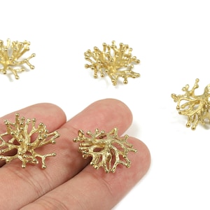 May include: Gold-colored, coral-shaped jewelry components. The components have a detailed, textured surface and a branching design. Several pieces are visible, some held in a hand, others scattered on a white background.