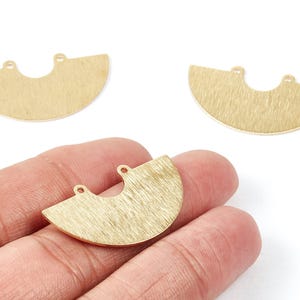 May include: Three gold-colored, half-circle shaped jewelry pendants. Each pendant has a textured surface and two small holes at the top for attaching to other jewelry components. One pendant is held in a hand.