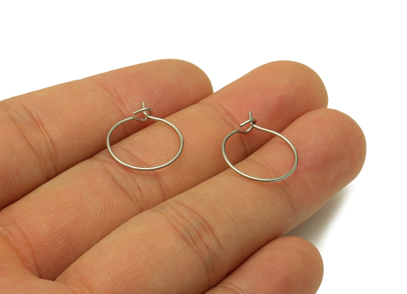 May include: Two silver hoop earrings with a hinged clasp. The earrings are shown on a person's hand.