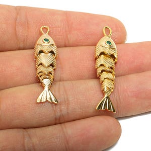 Könnte beinhalten: Zwei goldfarbene Anhänger in Fischform. Jeder Anhänger hat einen grünen Edelstein als Auge und eine Schlaufe zum Aufhängen. Die Fische haben detaillierte Schuppen und Flossen und sind wahrscheinlich Schmuckkomponenten.