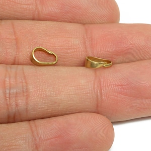 May include: Two small, gold-coloured jewellery bails are shown against a skin-toned background. One bail is oval-shaped, while the other is a curved, rectangular shape. These findings are used for jewellery making.