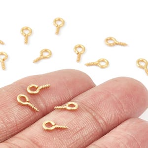 Può includere: Piccole viti a occhiello dorate. Questi elementi di fissaggio per la creazione di gioielli hanno un'estremità a vite e un occhiello ad anello per il fissaggio. I perni sono mostrati in una mano e sparsi su una superficie bianca.