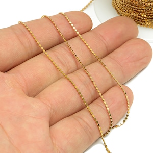May include: A close-up of a gold-colored chain with small, round links. The chain is lying on a hand, and the background is white.