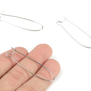 May include: Three silver-colored earring hooks are displayed against a white background. One hook is held by a hand. The hooks are oval-shaped with a looped top for attaching to the ear. The hooks are made of thin, shiny metal.
