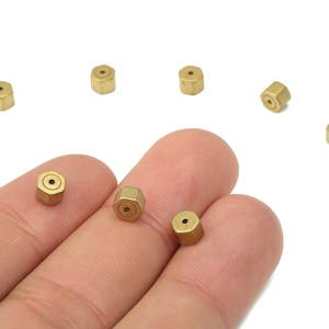 May include: A collection of small, gold-toned, hexagonal beads. Each bead has a central hole. The beads are scattered on a white surface, with some held in a hand. The beads are approximately 0.5 cm in diameter.