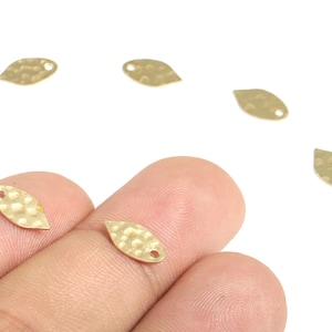 May include: Close-up of several small, gold-coloured leaf-shaped charms with a hammered texture and a single hole for stringing. Some charms are held by a person's fingers. These charms are ideal for jewellery making.