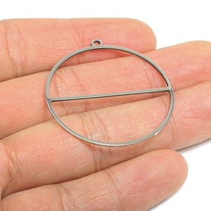 Könnte beinhalten: Ein silberfarbenes Metall-Ohrringteil mit kreisförmigem Design und einem horizontalen Balken in der Mitte. Das Teil hat oben eine kleine Schlaufe zur Befestigung. Schmuckherstellungszubehör.