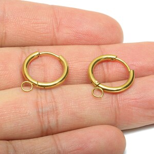 May include: Two gold-coloured hoop earrings with a hinged closure and small loop for attaching charms. The earrings are round and have a polished finish. The jewellery is displayed on a light background.