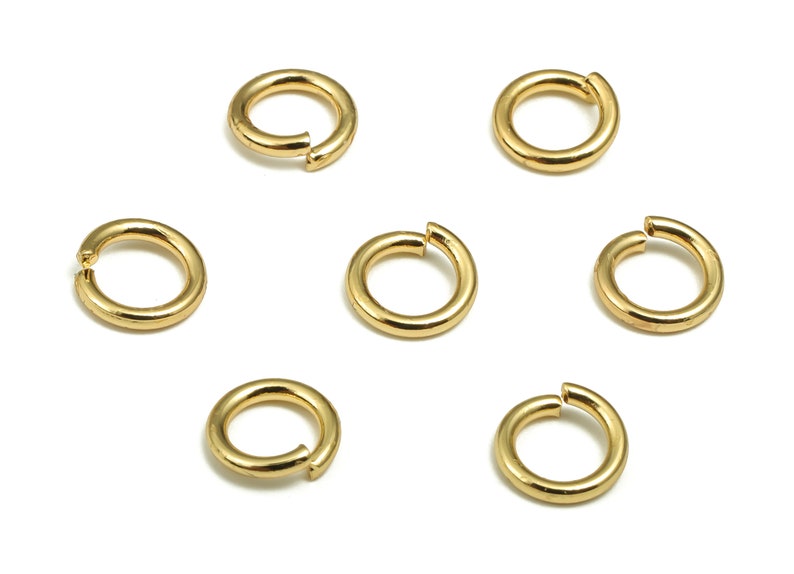 Op de afbeelding: Zeven gouden springringen, ook wel splitringen genoemd, zijn in een cirkelvormige opstelling op een witte achtergrond geplaatst. Deze ringen worden vaak gebruikt bij het maken van sieraden en handwerk.