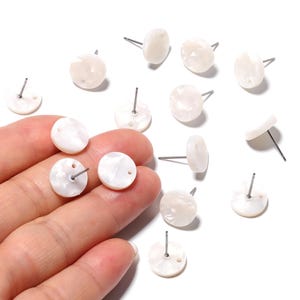 May include: A collection of white, round stud earrings with a pearlescent finish. Each earring has a silver post for piercing. Some earrings are held in a hand, while others are scattered on a white surface.
