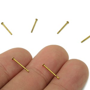 Op de afbeelding: Close-up van verschillende kleine, goudkleurige metalen pinnen met platte koppen. Twee pinnen worden tussen de vingers gehouden, terwijl andere erboven verspreid zijn. De pinnen staan op een witte achtergrond.