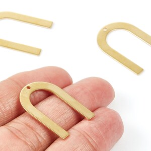 May include: Three gold-colored, U-shaped metal charms. One charm is held in a hand, while the other two are scattered above. Each charm has a small hole at the top, likely for stringing or attaching to other items. The charms have a matte finish.