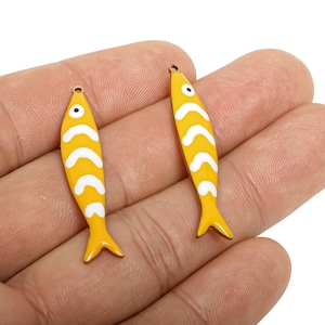 Può includere: Due ciondoli a forma di pesce giallo con motivi a onde bianche e piccoli dettagli degli occhi. I ciondoli hanno un piccolo foro nella parte superiore per l'infilatura. I ciondoli a forma di pesce sono di un giallo brillante con accenti bianchi e una piccola coda.