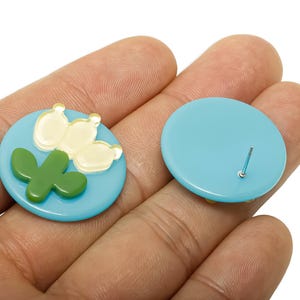 May include: A pair of light blue stud earrings. One earring features a green cactus and cream-coloured flowers on a blue disc. The other earring is a plain blue disc with a silver post. Jewellery.
