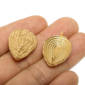 May include: Two gold-coloured stud earrings. One earring has a woven, textured design, while the other has a smooth, ridged surface. The earrings are held in a hand against a white background. The earrings have a post for piercing.