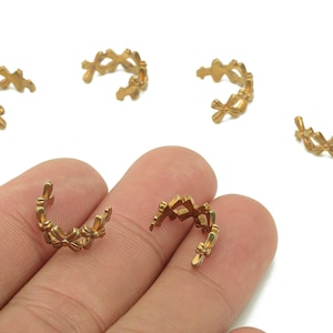 May include: Gold-colored metal ear cuffs with a repeating cross-like design. The ear cuffs are in a curved shape, and several are scattered on a white background. One is held in a hand.