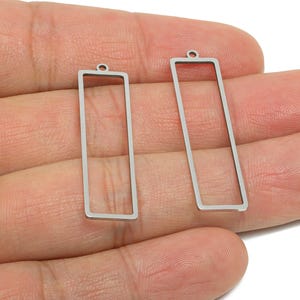 May include: Two silver-colored rectangular earring charms with a small loop at the top. The charms are held in a hand, showcasing their simple, open-frame design. These charms are suitable for jewelry making.