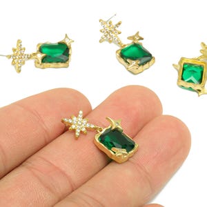 May include: Gold-toned earrings with a starburst and emerald-green rectangular stone. The earrings are dangling and have a post backing. One earring is held in a hand, while three others are scattered on a white surface.