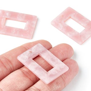 Può includere: Tre perline rettangolari rosa chiaro in acrilico con effetto marmorizzato e un ritaglio rettangolare. Una perlina è tenuta in mano. Le perline sono per la creazione di gioielli.