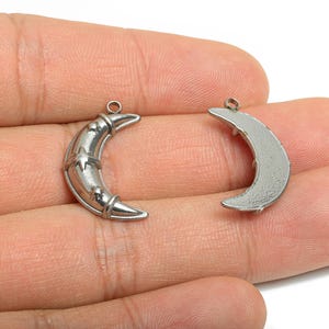 Op de afbeelding: Twee zilverkleurige maanvormige bedels. Eén bedel heeft een sterontwerp, de andere is effen. Beide hebben een kleine lus om aan sieraden te bevestigen. De bedels worden in een hand gehouden. De bedels zijn van metaal.