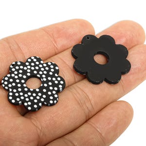 May include: Two black flower-shaped pendants. One has a white polka dot pattern, the other is solid black. Both have a hole in the centre and a small hole at the top for hanging. Likely jewellery components.