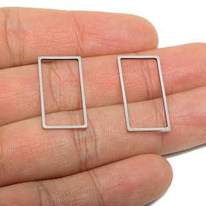 Puede incluir: Dos componentes de pendientes rectangulares de color plateado. Cada componente es un rectángulo fino y abierto. Los pendientes se muestran sobre un fondo color piel. Estos componentes son adecuados para la fabricación de joyas.