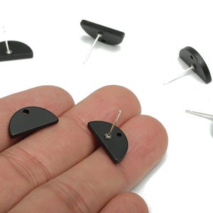Puede incluir: Pendientes de botón negros en forma de semicírculo con postes plateados. Los pendientes están hechos de un material negro mate y tienen un pequeño agujero en la parte superior. Varios pendientes están dispersos sobre un fondo blanco.