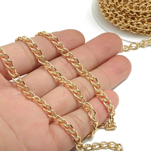 May include: A gold chain with a curb link pattern. The chain is shown in a hand, with a spool of the chain in the background.